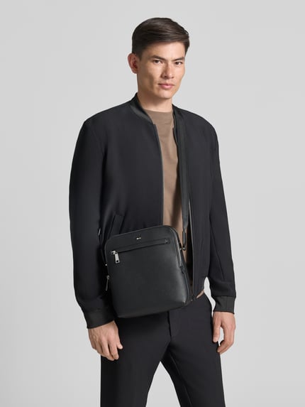 Mężczyzna nosi Umhängetasche mit Label-Prägung Modell 'RAY_BIG NS' od BOSS w czarnej skórzanej optyce, główna komora, pasek na ramię, sztuczna skóra, widok całej sylwetki z przodu.