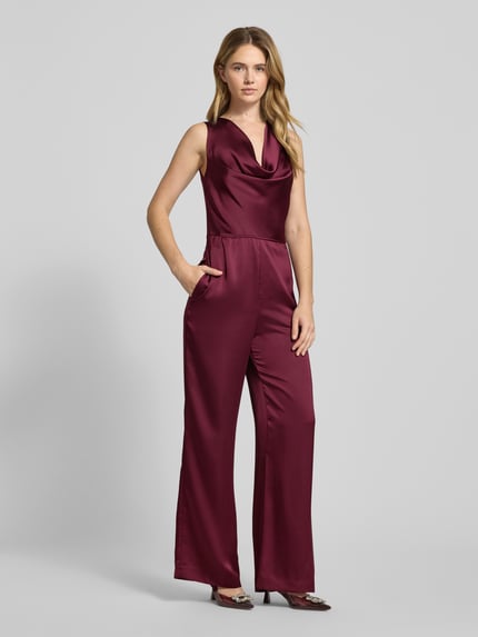 Modelka kobieca nosi bordowy, taliowany kombinezon z satyny Taillierter Jumpsuit aus Satin Modell 'KIMARI-1' od HUGO, jednolity kolor, aplikacja w formie kokardy, dekolt wodospadowy, kryte zamki, całopostaciowy widok z przodu.