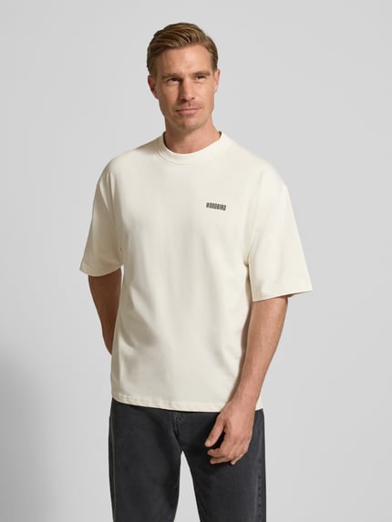 Männermodel trägt T-Shirt mit Label-Print Modell 'Beam' von WOODBIRD in Creme, lockerer Schnitt, reine Baumwolle, Nahaufnahme Oberkörper.