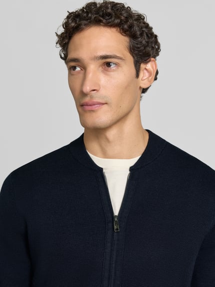 Männermodel trägt Slim Fit Strickjacke aus Baumwoll-Mix mit Stehkragen von Christian Berg Men, dunkle Farbe, Nahaufnahme Oberkörper. Kontext: Herren Strickjacke, Zweiwege-Reißverschluss, langärmliges Design, gerippte Abschlüsse.
