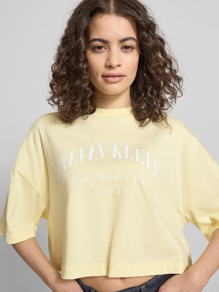 Vrouwenmodel draagt Cropped T-Shirt met Logo-Print van Calvin Klein Jeans in lichtgeel, puur katoen, korte fit, ronde hals, ruglengte maat S 42 cm, logoprint, Nahaufnahme bovenlichaam.