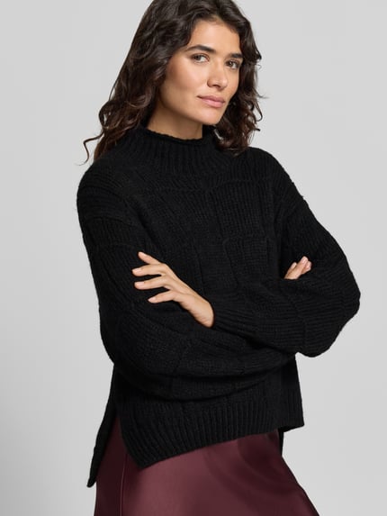 Modelka nosi sweter wełniany z strukturą, jednokolorowy, imitacja dzianiny, krótki krój, szwy działowe, kołnierz typu mock neck, długie rękawy z prążkowanymi zakończeniami, wykończenie dołu prążkowane, 'Pullover aus Wolle mit Strukturstrick' od MARC CAIN, pudełkowy krój, widok całej sylwetki z przodu.