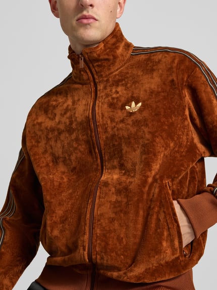 Männermodel trägt Sweatjacke mit Samtoptik Modell 'FIREBIRD TT VEL' von adidas Originals, braun glänzend, Logo-Print, Nahaufnahme Oberkörper.