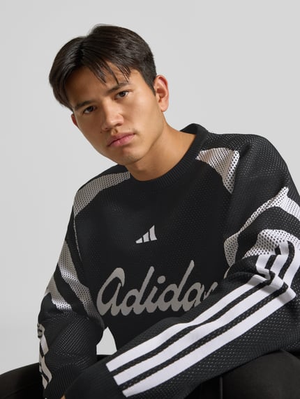 Mannelijk model draagt Pullover met groot logo-opschrift van adidas Sportswear, meerkleurige gebreide look, strepen, logo details, ronde hals, geribde boorden, motiefprint, nahaufnahme bovenlichaam.