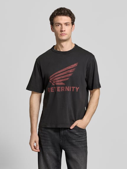 Męski model nosi T-Shirt mit Label-Print Modell 'AÉRO' od RETERNITY, czarna bawełna, krój regular fit, okrągły dekolt, długość tyłu 67 cm w rozmiarze M, widok całej sylwetki z przodu.