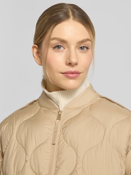 Kobiece model prezentuje kurtkę pikowaną z zamkiem błyskawicznym dwustronnym model 'IMANE' od Beaumont Amsterdam w kolorze beżowym, krój relaxed fit, stójka, damska, szerokość ramion w rozmiarze 36: 44 cm, szerokość klatki piersiowej w rozmiarze 36: 60 cm, długość tyłu w rozmiarze 36: 55 cm, długość rękawów w rozmiarze 36: 61 cm, zbliżenie na górną część ciała.