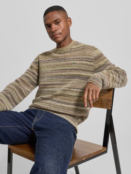 Mannelijk model draagt Regular Fit gebreide pullover uit wolmix van Lacoste in gemêleerde look met labelpatch, lange mouw, ronde hals, rompzoom, knitwear, zittend op stoel, Nahaufnahme Oberkörper.