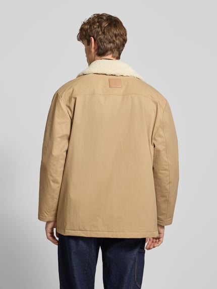 Männermodel draagt Steppmantel mit Label-Print van Calvin Klein Jeans in beige, rechte vormen, zachte textuur, Ganzkörperansicht Rückansicht, regular fit, capuchon met tunnelkoord, drukknopzakken, verborgen ritssluiting, ruglengte 80 cm, schouderbreedte 53 cm, borstwijdte 64 cm, mouwlengte 64 cm.
