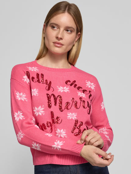Frauenmodel trägt Pullover mit Pailletten-Schriftzug von VERO MODA in Pink mit weißem Sternenmuster und Glitzer-Print, Rundhals, Strickoptik, mehrfarbig, Nahaufnahme Oberkörper.