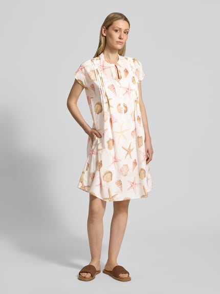Vrouwenmodel draagt Knielanges Kleid mit Tunikakragen van Emily Van den Bergh, puur katoen, getailleerd model, knielengte, tuniekkraag, biezen, korte knoopsluiting, ruglengte bij maat 36: 96 cm, borstwijdte bij maat 36: 54 cm, wit met schelp- en zeesterprint, zachte textuur, volledige lichaamsopname van voren.