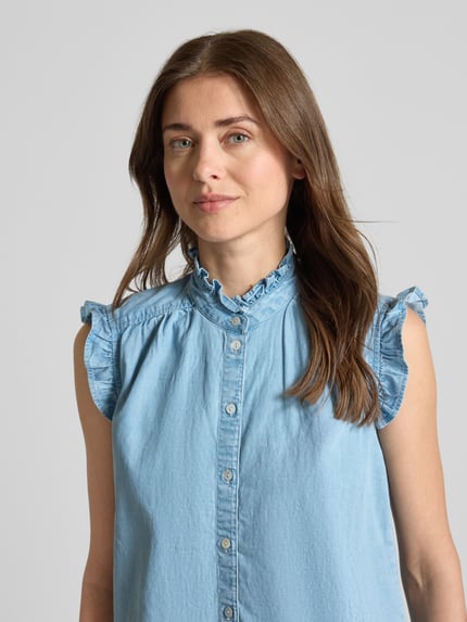 Modelka kobieca nosi Blusentop in Denim-Optik mit Rüschen od Christian Berg Woman, mieszanka lyocellu i bawełny, krój regular fit, tkanina stylizowana na denim, stójka, falbany, długość tyłu w rozmiarze 36: 57 cm, długość rękawów w rozmiarze 36: 50 cm, jasny niebieski kolor, prosta faktura, detaliczne guziki, widok z przodu, zbliżenie na górną część ciała.