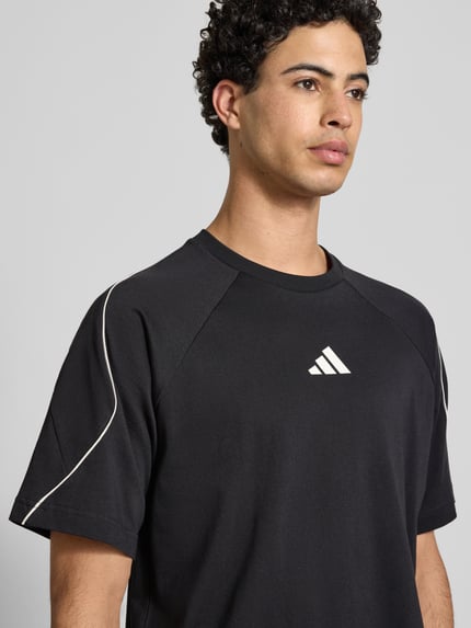 Männermodel trägt T-Shirt mit Label-Print von adidas Sportswear in Schwarz, reine Baumwolle, Regular-Fit, gerippter Rundhalsausschnitt, Nahaufnahme Oberkörper.
