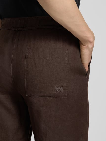 Frauenmodel trägt Leinenhose mit Bindegürtel Modell 'Chiara' von MAC, dunkelbrauner Farbton, sichtbare Struktur, Nahaufnahme Rückansicht, reines Leinen, französische Eingrifftaschen, elastischer Bund.