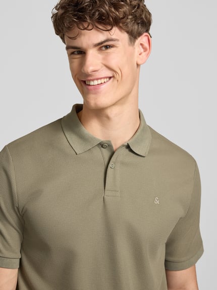 Męski model nosi Regular Fit Poloshirt mit Strukturmuster Modell 'AUSTIN' von Jack & Jones, kolor khaki, bawełniany materiał, gładka faktura, krótki rękaw, krój regular fit, guzikowa plisa, stitching, widoczne logo, zdjęcie: zbliżenie na górną część ciała.