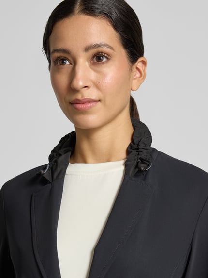 Frauenmodel trägt Slim Fit Blazer mit Stretch-Anteil von Marc Cain, Schwarz, Reverskragen mit Tunnelzug, Nahaufnahme Oberkörper. Elastischer Material-Mix, Knopfleiste, Reißverschlusstaschen, Rückenlänge 25 cm, Brustweite 43 cm, Ärmellänge 63 cm.