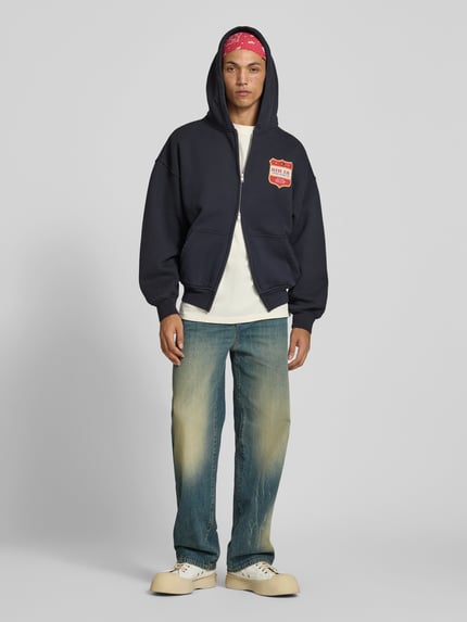 Männermodel trägt Oversized Sweatjacke aus Baumwolle von RHUDE in Dunkelblau mit Label-Print, Zweiwege-Reißverschluss und Känguru-Tasche, reine Baumwolle, Ganzkörperansicht von vorne.