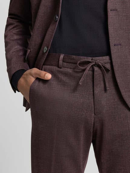 Męski model nosi spodnie materiałowe Straight Fit Stoffhose mit Tunnelzug Modell 'Sparta' von Zuitable, bordowy, kieszenie wpuszczane, zakładki w pasie, szlufki na pasek, szerokość w rozmiarze 50: 43 cm, długość wewnętrzna nogawki 82 cm, zbliżenie na pas