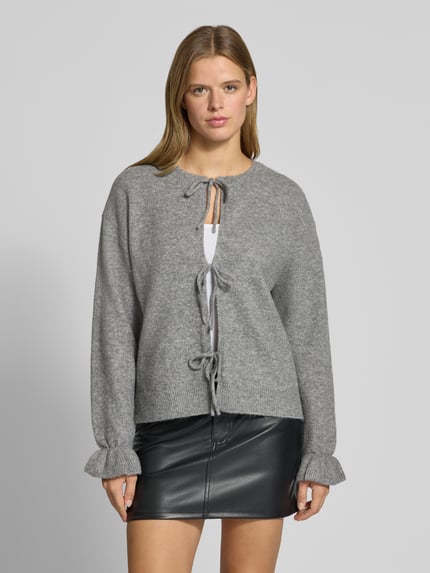 Frauenmodel trägt Strickjacke mit Schleifenverschluss Modell 'PCSALLY' von PIECES in Grau, Melange-Optik, strukturierte Stricktextur, Regular Fit, cropped, Schnürung, überschnittene Schultern, ausgestellte Ärmel, gerippter Rumpfabschluss, Ganzkörperansicht von vorne.