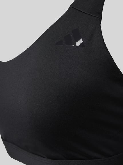 Frauenmodel trägt Sport-BH mit Logo-Detail von adidas Sportswear in Schwarz aus Elastischer-Material-Mix, eng anliegender Schnitt, Nahaufnahme Oberkörper.