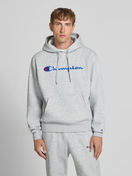 Männermodel trägt Hoodie mit Label-Stitching von Champion in Hellgrau, Baumwoll-Mix, lockerer Schnitt, Ganzkörperansicht von vorne.