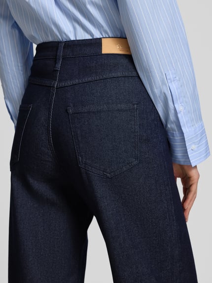 Frauenmodel trägt Wide Leg Jeans aus Viskose-Baumwoll-Mix Modell 'SURI' von s.Oliver BLACK LABEL, stylizowana na denim, naszywka z logo, nity, 5 kieszeni, szlufki na pasek, zapięcie na guzik i zamek błyskawiczny, mieszanka wiskozy i bawełny, krój wide fit, wysoki stan, skrócony krój, Ganzkörperansicht Rückansicht
