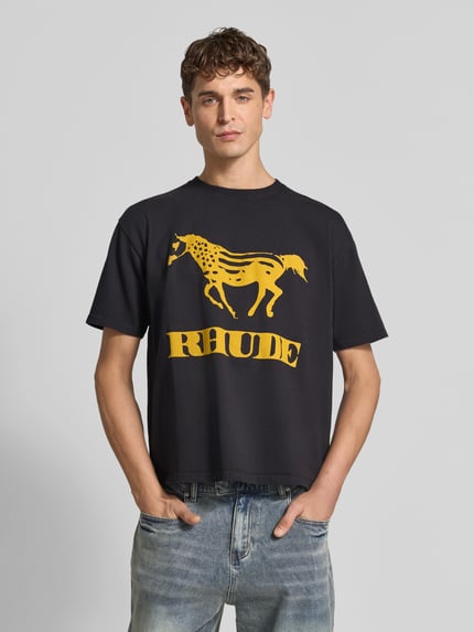 Männermodel trägt T-Shirt mit Label-Print von RHUDE, schwarze Baumwolle, Rundhalsausschnitt, gelber Print, gerader Schnitt, Ganzkörperansicht von vorne.
