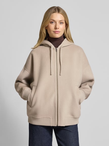 Frauenmodel trägt Oversized Sweatjacke mit Kapuze Modell 'DUNSTONE' von Drykorn im Woll-Mix, beige, Reißverschluss, Eingrifftaschen, gerippte Abschlüsse, Ganzkörperansicht von vorne.