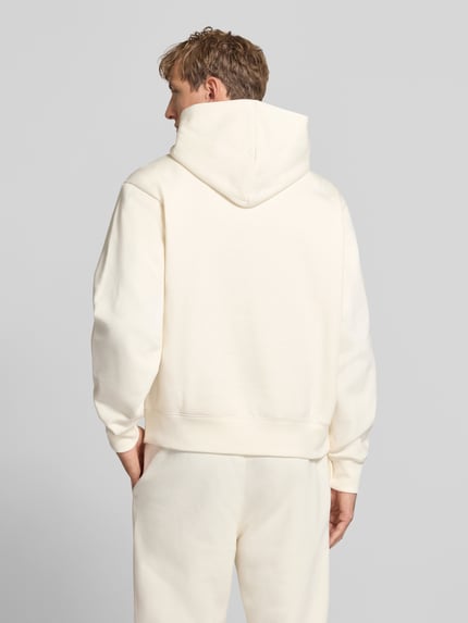 Männermodel trägt Hoodie mit Label-Stitching von Champion aus Baumwoll-Mix, lockerer Schnitt, cremefarben, Ganzkörperansicht Rückansicht.