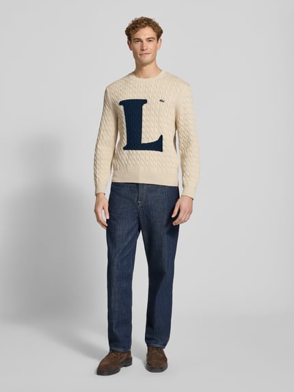 Männermodel trägt Regular Fit Strickpullover aus Baumwoll-Mix von Lacoste in Beige mit großem blauem L, kombiniert mit Jeans, Ganzkörperansicht von vorne. Herren Strickjacke von Lacoste.