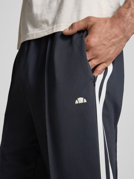Männermodel trägt Straight Fit Sweatpants mit Logo-Detail Modell 'Latucci' van Ellesse in donkerblauw met witte strepen, elastische tailleband, siernaden, detailopname rechterbeen, context: herensweatpants elastische materiaalmix, binnenbeenlengte maat M 79 cm.