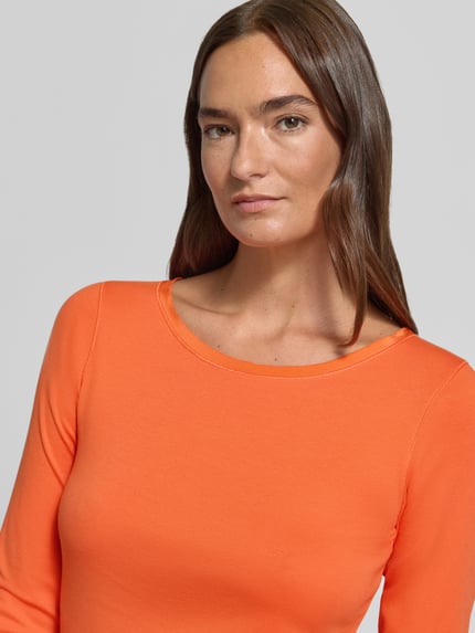 Frauenmodel trägt Slim Fit Langarmshirt aus Baumwoll-Mix von Marc Cain in Orange, U-Boot-Ausschnitt, Logo-Stickerei, Nahaufnahme Oberkörper.