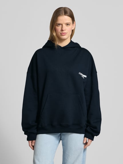 Frauenmodel trägt Oversized Hoodie mit Label-Stitching Modell 'No Hustle' von Karo Kauer in Dunkelblau, Baumwoll-Mix, Känguru-Tasche, Kapuze, elastische Abschlüsse, rückseitiger Statement-Print, Oversized-Fit, Ganzkörperansicht von vorne.