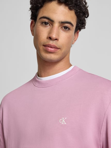Männermodel trägt Sweatshirt aus reiner Baumwolle von Calvin Klein Jeans in Rosa mit Label-Stitching, Nahaufnahme Oberkörper.