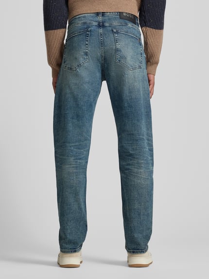 Männermodel trägt Tapered-Fit Jeans aus Baumwoll-Mix Modell 'TROY' von BOSS Orange in Denim-Optik mit Label-Patch, Nieten, Destroyed-Details, 5-Pocket-Design; Ganzkörperansicht Rückansicht.