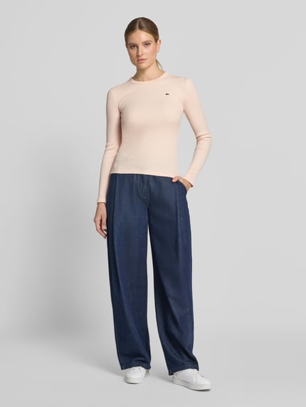 Frauenmodel trägt Slim Fit Langarmshirt aus reiner Baumwolle von Lacoste in Beige zu dunkelblauer Hose, Damen T-Shirt von Lacoste, Ganzkörperansicht von vorne.