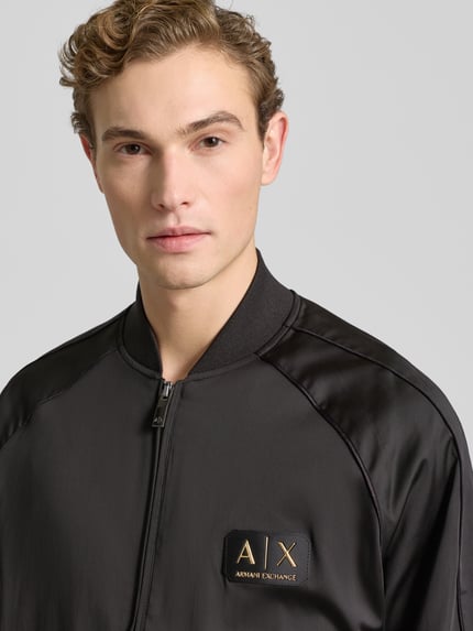 Męski model nosi Bomberjacke mit Reißverschluss od ARMANI EXCHANGE, czarny materiał z bawełną, zamek, detal z logo, zbliżenie na górę ciała.