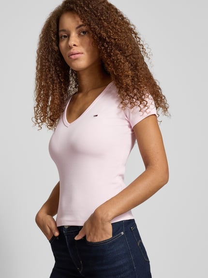 Modelka kobieca nosi Slim Fit T-Shirt aus Baumwoll-Mix od Tommy Jeans, jasnoróżowy, prążkowany, krótki rękaw, logo, widok z przodu w półzbliżeniu.