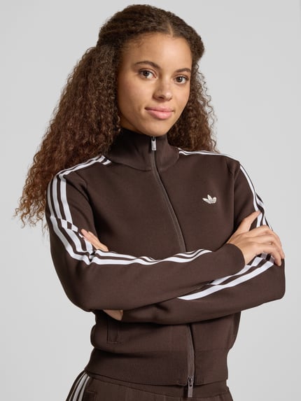 Frauenmodel trägt Strickjacke mit Zweiwege Reißverschluss von adidas Originals, braun, Rippstrick, Logo, Kontraststreifen, Nahaufnahme Oberkörper.