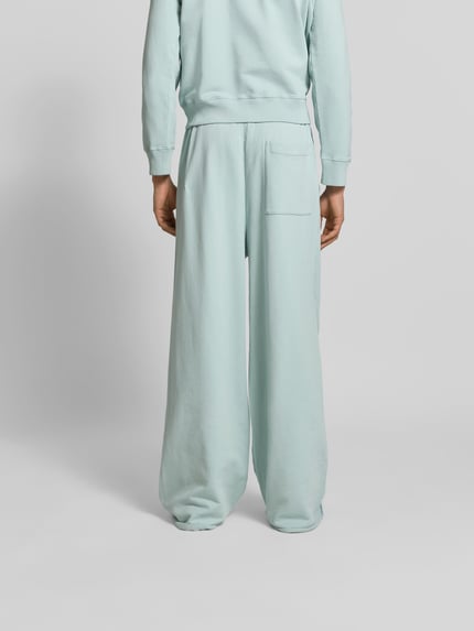 Męski model nosi Wide Fit Sweatpants mit elastischem Bund von MM6 Maison Margiela z bawełny, wyhaftowane logo, kieszeń z tyłu, kolor jasnoniebieski, krój wide fit, widok całej sylwetki z tyłu.