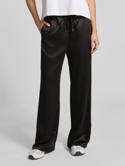 Kobieta nosi Wide Leg Stoffhose aus Satin od Juvia w kolorze czarnym, satynowa tekstura, luźny krój, elastyczny pas ze sznurkiem, kieszenie, szerokość w rozmiarze S: 40 cm, długość wewnętrzna nogawki: 80 cm, widok całej sylwetki z przodu