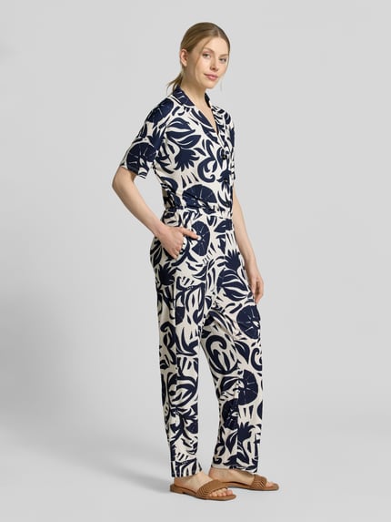 Frauenmodel trägt Jumpsuit mit floralem Allover-Print von Oui, Elastischer-Material-Mix, Taillierte Passform, Reverskragen, Kurzärmelig, Knopfleiste, Grundfarben Creme und Dunkelblau, weiche strukturierte Oberfläche, Ganzkörperansicht von vorne. Rückenlänge bei Größe 36: 146 cm, Brustweite: 51 cm, Ärmellänge: 13 cm, Gesamtlänge: 146 cm, Innenbeinlänge: 76 cm.