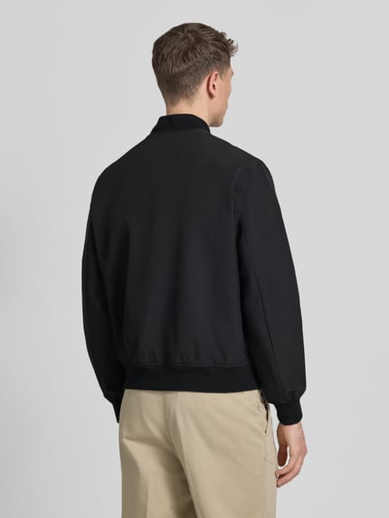 Męski model nosi bomberkę z mieszanki materiałów z dodatkiem streczu Jack & Jones Premium, model 'BLAASPEN', kolor czarny, luźny krój, stójka, zamek błyskawiczny, wpuszczane kieszenie boczne, elastyczne wykończenia, długość tyłu w rozmiarze M: 65 cm, szerokość ramion w rozmiarze M: 48 cm, szerokość klatki piersiowej w rozmiarze M: 60 cm, długość rękawów w rozmiarze M: 66 cm, widok całej sylwetki z tyłu.
