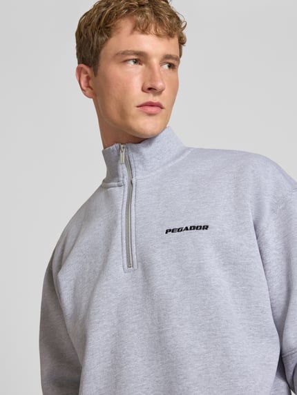 Männermodel trägt Relaxed Fit Sweatshirt aus Baumwoll-Mix von PEGADOR in Hellgrau mit Troyer-Kragen, Logo-Stickerei, Nahaufnahme Oberkörper.
