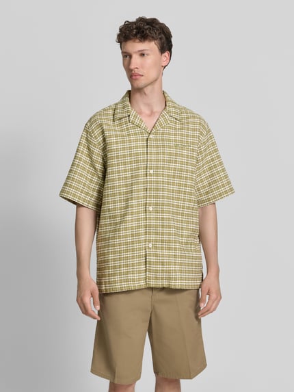 Männermodel trägt Regular Fit Freizeithemd aus reiner Baumwolle von Carhartt Work In Progress mit Strukturmuster, Halbarm, Durchgehende Knopfleiste, Brand-Stitching, Beige- und Grüntöne, Ganzkörperansicht von vorne.