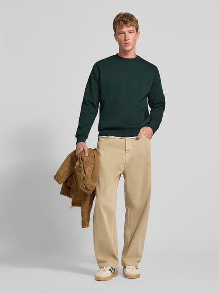 Männermodel trägt Regular Fit Sweatshirt mit Baumwoll-Anteil Modell 'Bradley' von JACK & JONES in Dunkelgrün, Rundhalsausschnitt, beige Hose, Ganzkörperansicht von vorne.