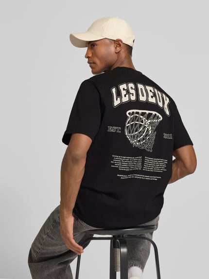 Männermodel trägt T-Shirt mit Label-Print Modell 'Brady whop' von Les Deux in Schwarz, Reine Baumwolle, Gerader-Schnitt, Rückenlänge 72 cm, Ganzkörperansicht Rückansicht.