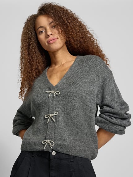 Frauenmodel trägt Pullover mit Schleifen-Schnürung von ONLY in Grau, Strukturstrick, Loose Fit, V-Ausschnitt, Ganzkörperansicht von vorne.