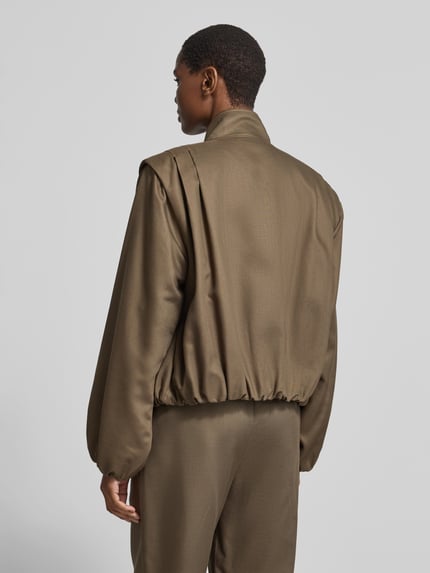 Frauenmodel trägt Funktionsjacke mit großen Fronttaschen von black palms in Uni-Oliv, strukturierte Webware mit elastischem Bund, Ganzkörperansicht Rückansicht.