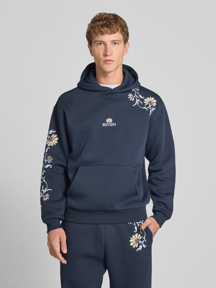 Männermodel trägt Relaxed Fit Hoodie mit Känguru-Tasche von REVIEW in Dunkelblau mit floraler Stickerei, Baumwoll-Mix, Ganzkörperansicht von vorne.