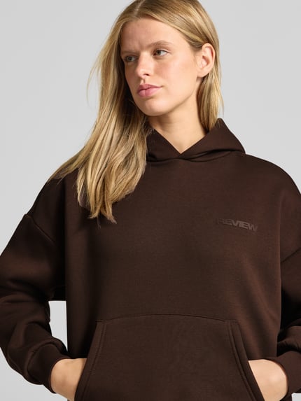 Frauenmodel trägt Relaxed Fit Hoodie aus Baumwoll-Mix mit Label-Detail von REVIEW, dunkle Farbe, Känguru-Tasche, elastische Abschlüsse, Nahaufnahme Oberkörper